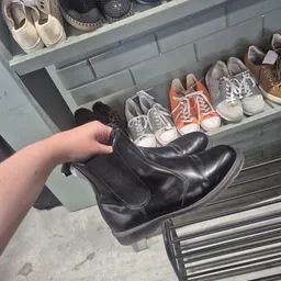 Hand toont zwarte laarzen boven uitgestalde schoenen in een grijze stellingkast