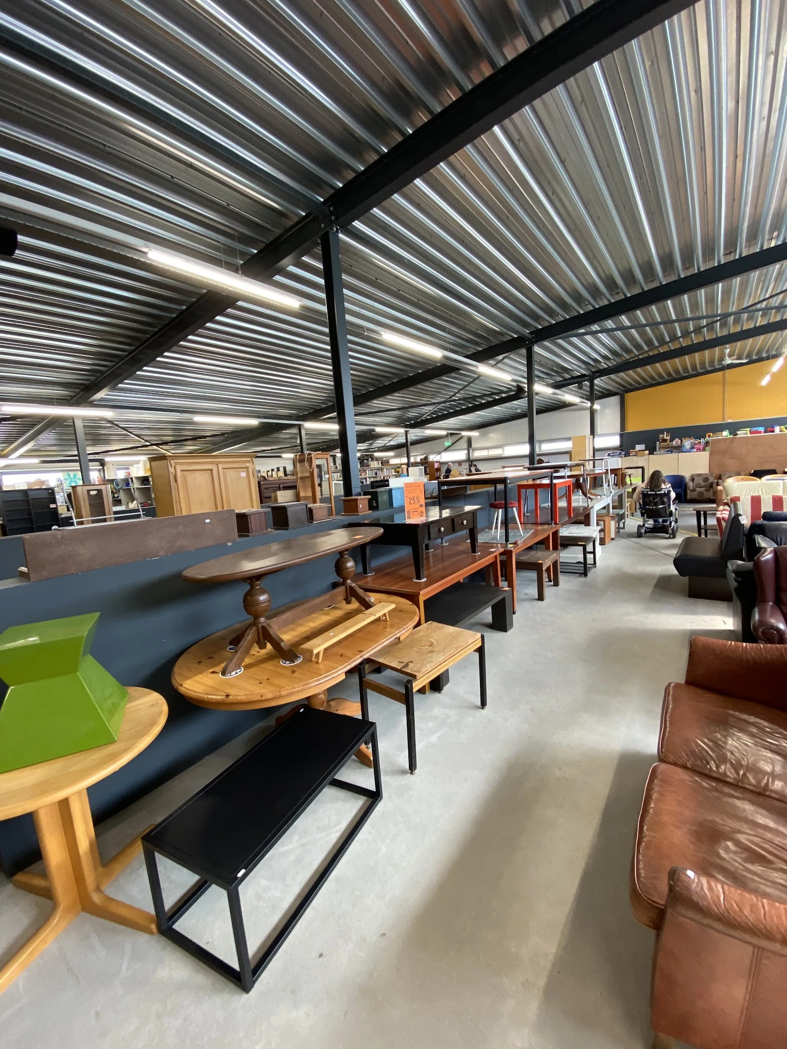 Winkelvloer van kringloopwinkel met houten meubels, tafels en stoelen op grijze betonvloer onder golfplaten dak