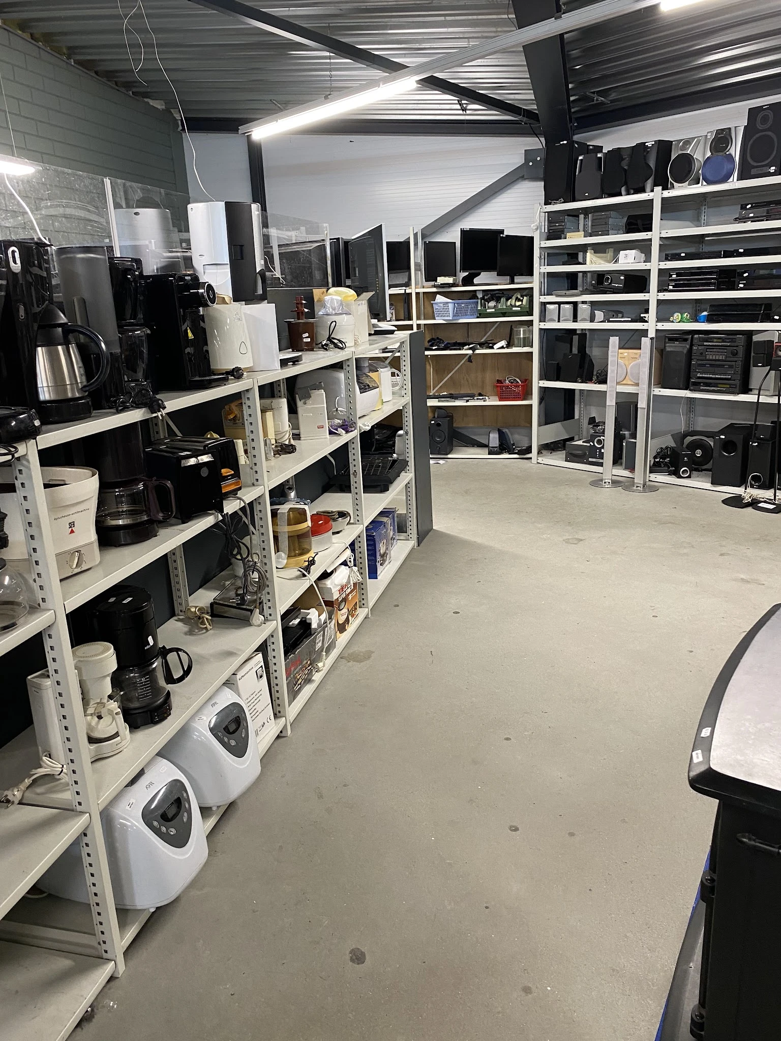 Wit stellingwerk met elektronische apparaten, huishoudelijke items en dozen in een opslagruimte van een kringloopwinkel