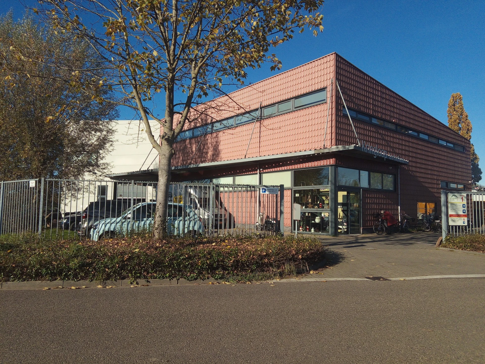 Voorzijde van kringloopcentrum met rode bakstenen gevel, grote ramen en ingang, parkeerplaats ervoor