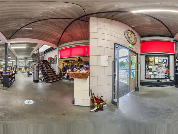 Panoramisch overzicht van het interieur van een kringloopwinkel met trap, stellingen, toonbank en rode signage