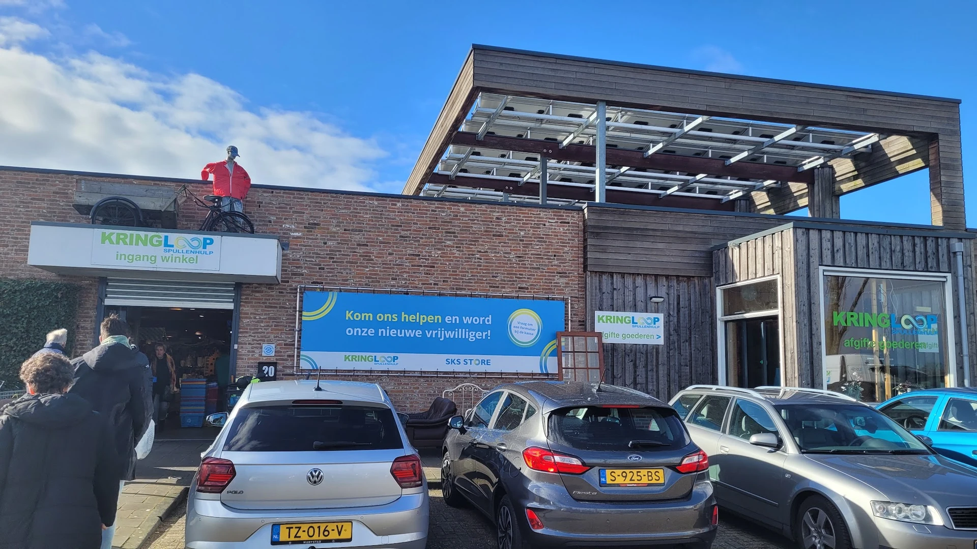 Voorgevel van kringloopwinkel met blauwe banner, parkeerplaatsen en moderne uitbouw