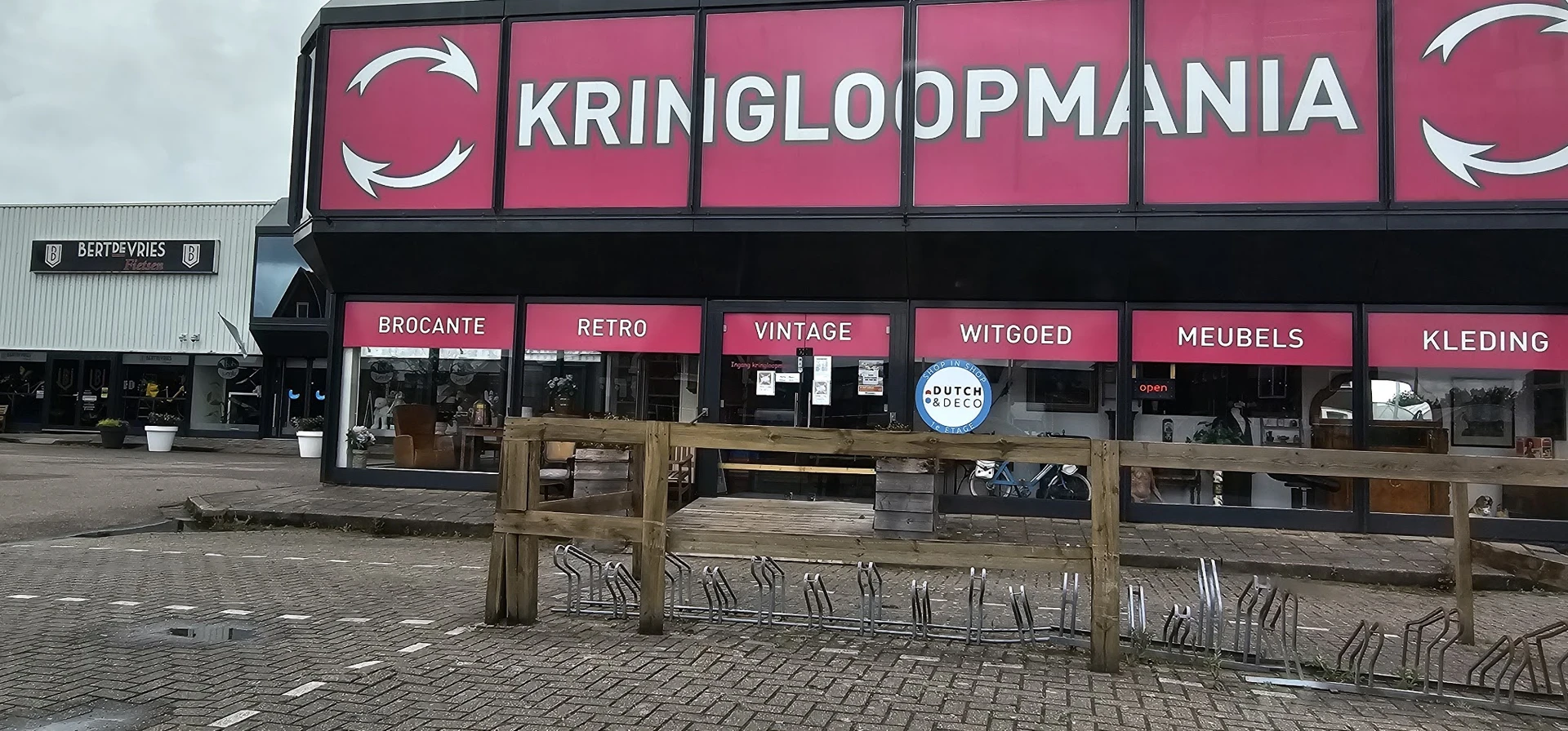 Gevel van Kringloopmania met roze uithangbord en houten terras ervoor met verschillende categorieën
