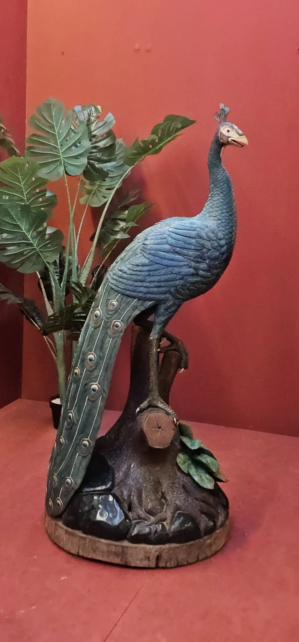 Blauw decoratief vogelfiguur met lange staart en groene bladeren tegen rode achtergrond