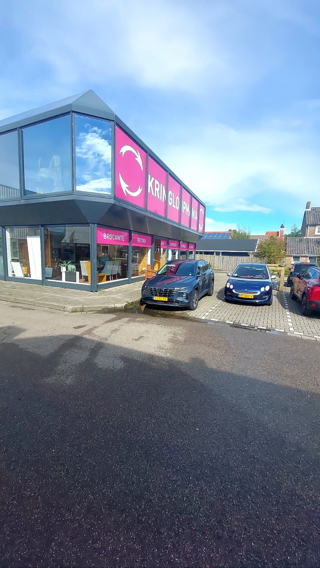 Voorkant van kringloopwinkel met roze gevel en parkeerplaats met auto's