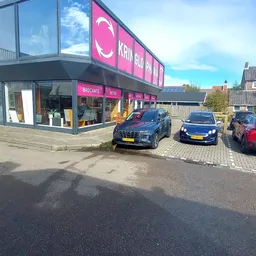 Voorkant van kringloopwinkel met roze gevel en parkeerplaats met auto's