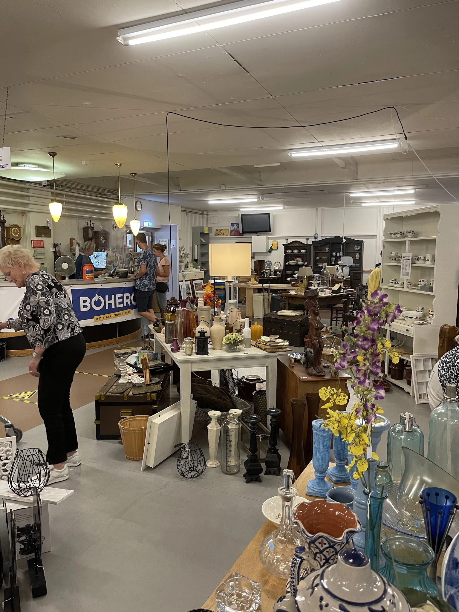 Winkelvloer van kringloopwinkel met diverse gebruikte items: vazen, bloemen, lampen, meubels en huishoudwaren op tafels en st