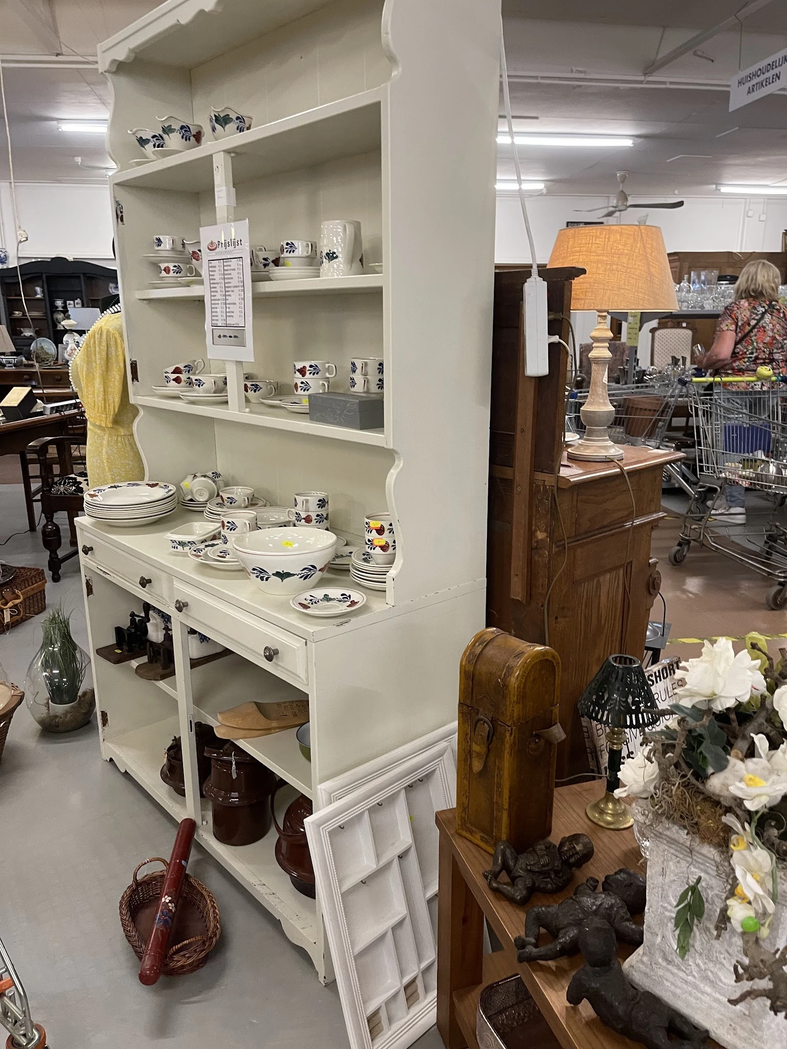 Witte dressoirkast met uitgestald serviesgoed, borden en decoratieve items in een kringloopwinkel