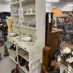 Witte dressoirkast met uitgestald serviesgoed, borden en decoratieve items in een kringloopwinkel