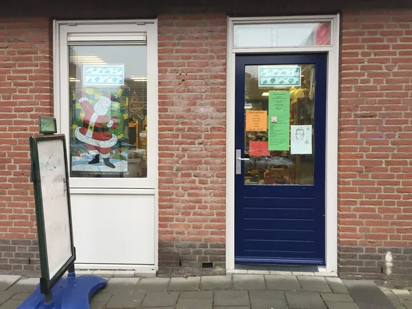 Voorgevel van kringloopwinkel met twee deuren, rode bakstenen muur, etalageruiten met kerstdecoratie en informatieborden
