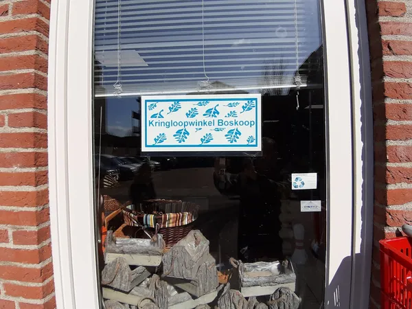 Winkelruit met bord 'Kringloopwinkel Boskoop' en uitzicht op tweedehands textiel en huishoudartikelen in de etalage
