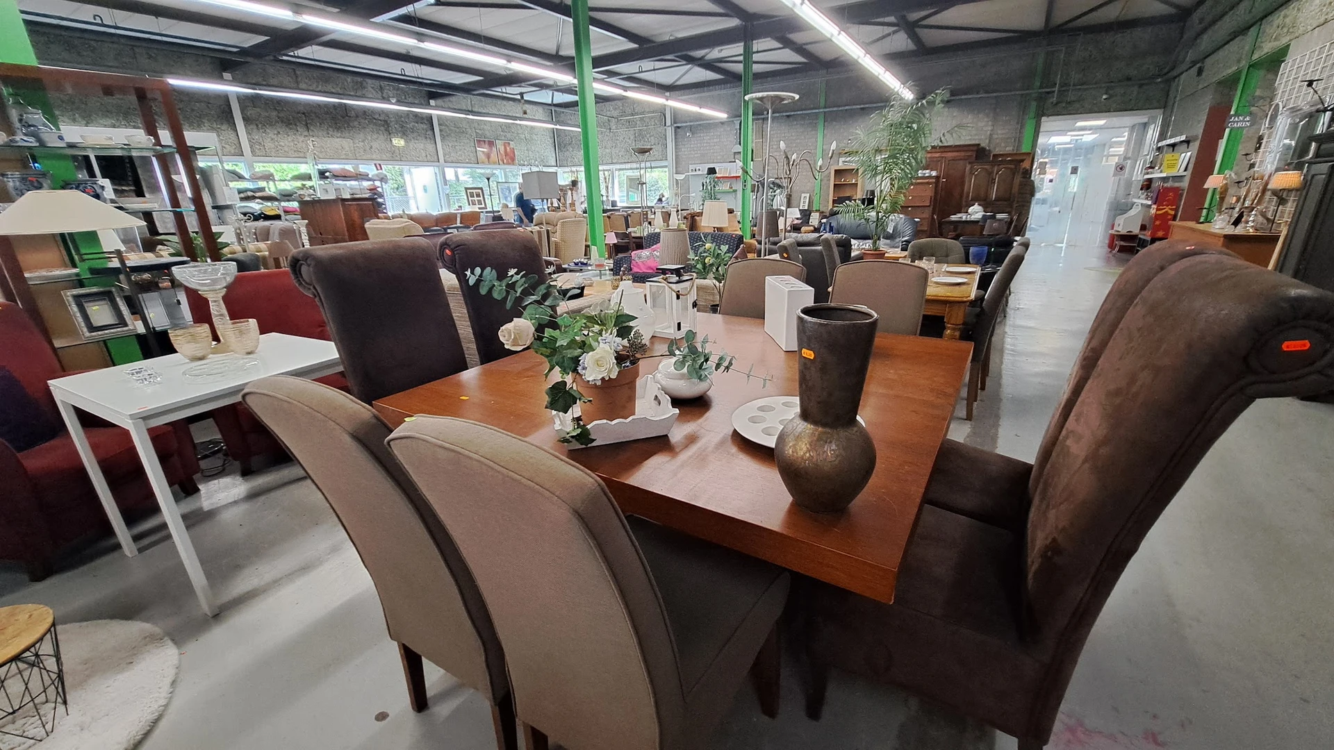 Houten eetdutafel met bruine stoelen en decoratieve vazen in een kringloopwinkel met groene zuilen en veel meubels op de acht