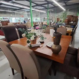 Houten eetdutafel met bruine stoelen en decoratieve vazen in een kringloopwinkel met groene zuilen en veel meubels op de acht
