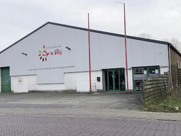Witte gevel van kringloopwinkel met rood logo 'Op 'e Nij' en rode deuren, aan straat met parkeerplaats