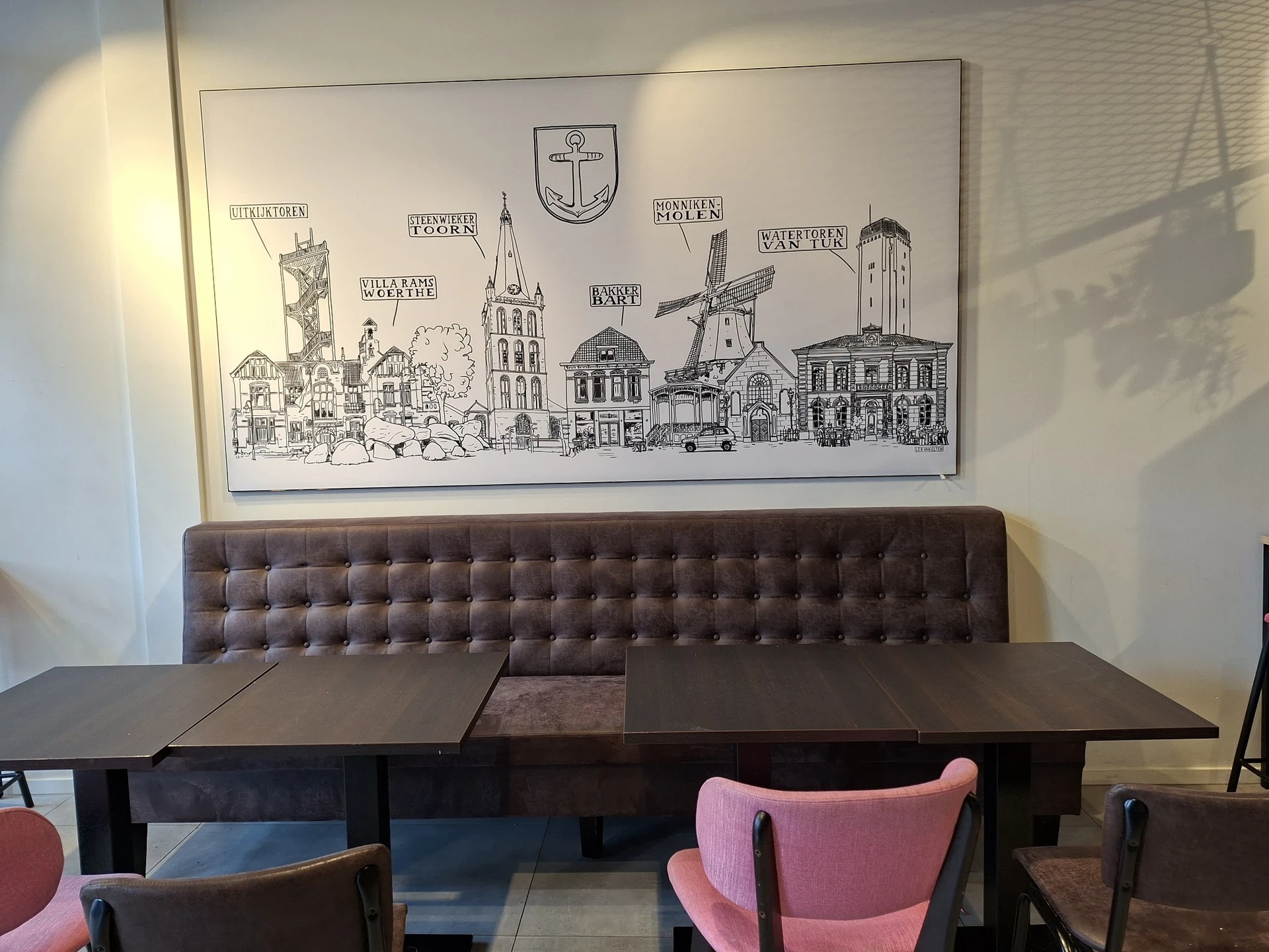 Zithoek met bruine gestoffeerde bank en roze stoelen aan zwarte tafels, onder decoratieve wandtekening van stadsgezicht