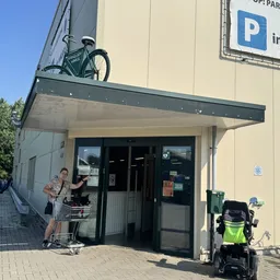 Ingang van kringloopwinkel met luifel, fietsen op het dak en daarnaast, persoon met winkelwagen
