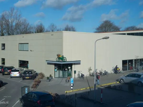 Voorkant van kringloopwinkel met witte gevel, ingang met groene bovenkant en fietsen ervoor, parkeerplaats met auto's