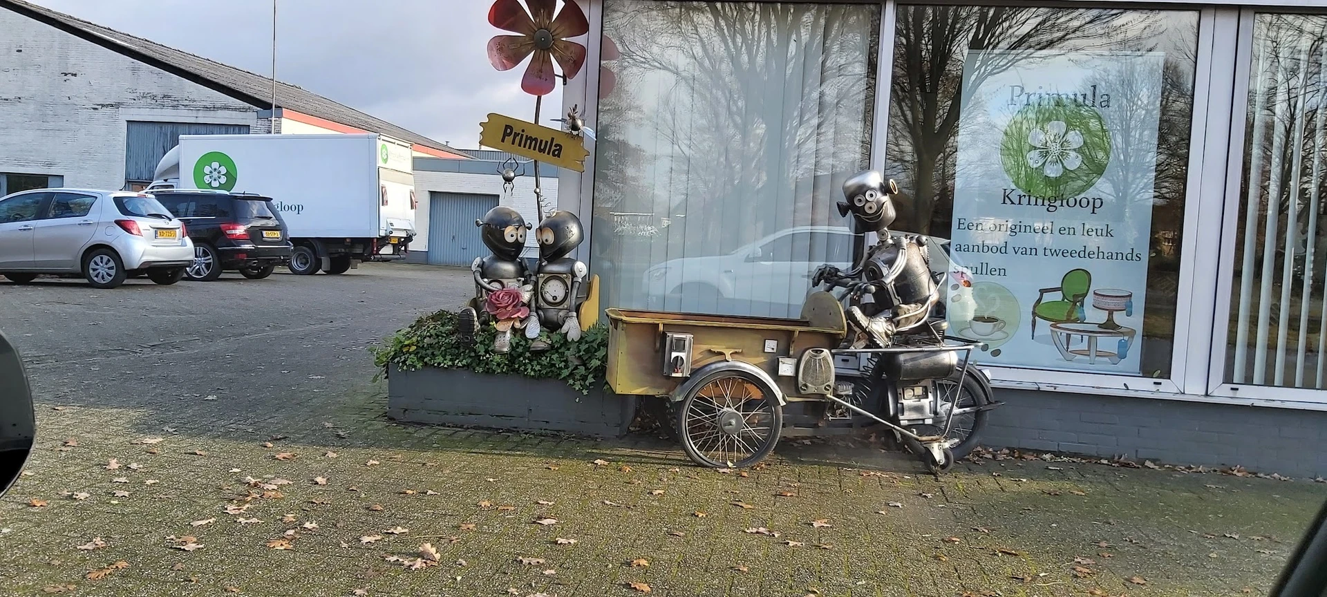 Decoratieve robot-figuren op een gele aanhanger naast de winkel, met winkellogo Primula zichtbaar