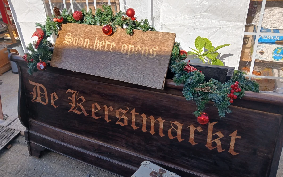 Zwarte houten plantersbak met gouden letters 'De Kerstmarkt' en aankondigingsbord 'Soon here opens' met kerstdecoratie buiten