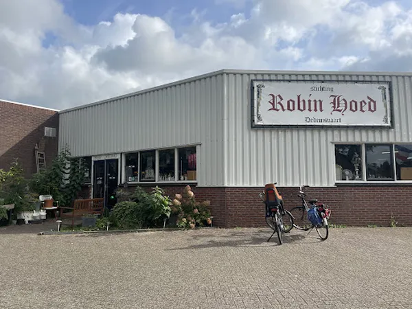 Voorkant van kringloopwinkel met naam Robin Hoed op gevel, ingang met ramen en fietsen ervoor