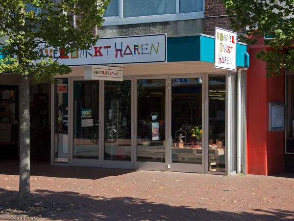 Gevel van kringloopwinkel met glazen deuren, winkelnaambord en groene klimplanten aan zijkanten