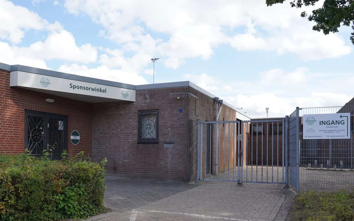 Voorgevel van bakstenen kringloopwinkel met 'Sponsorwinkel' tekst, ingang met blauwe hek en parkeerterrein