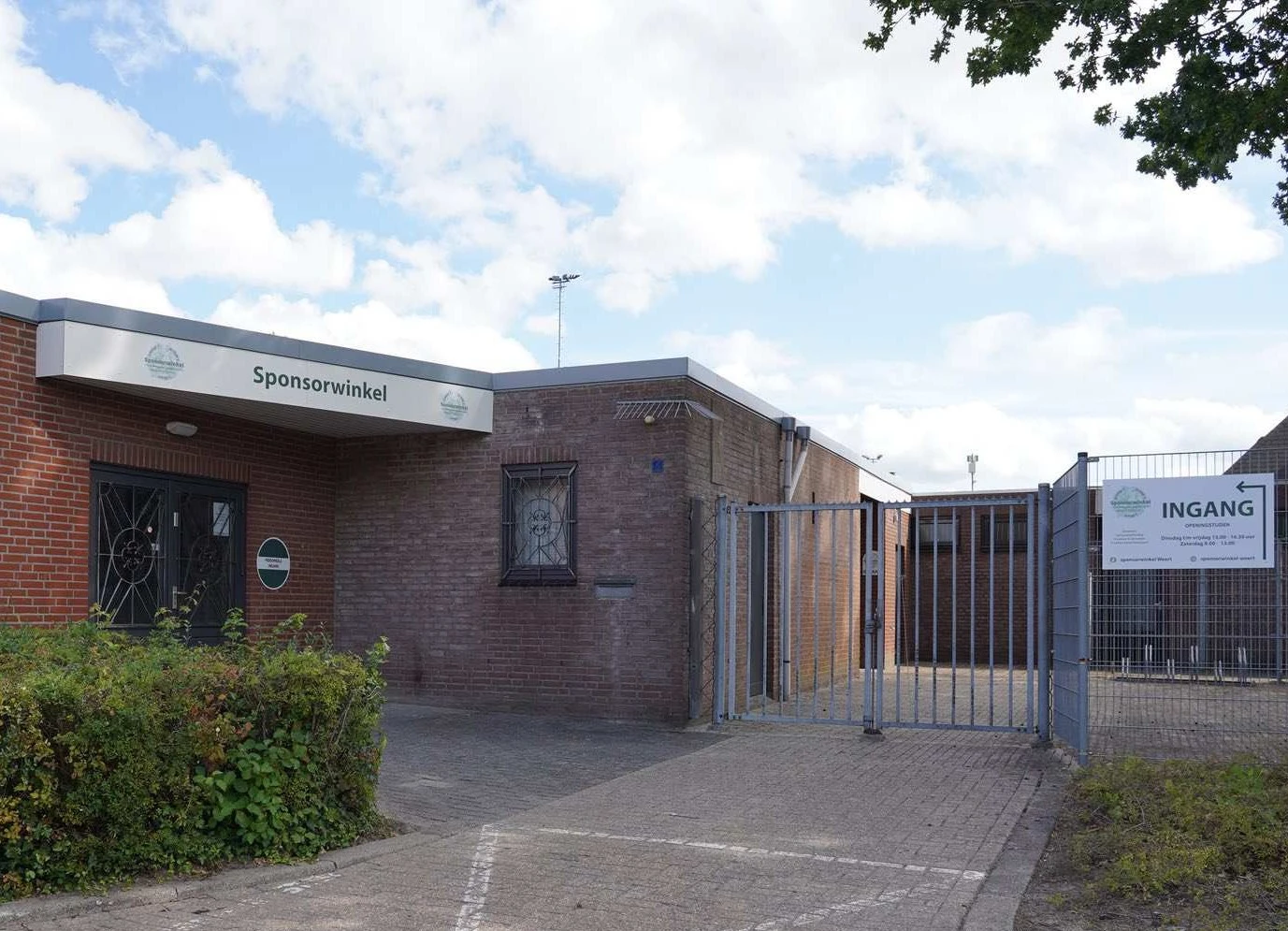 Voorgevel van bakstenen kringloopwinkel met 'Sponsorwinkel' tekst, ingang met blauwe hek en parkeerterrein