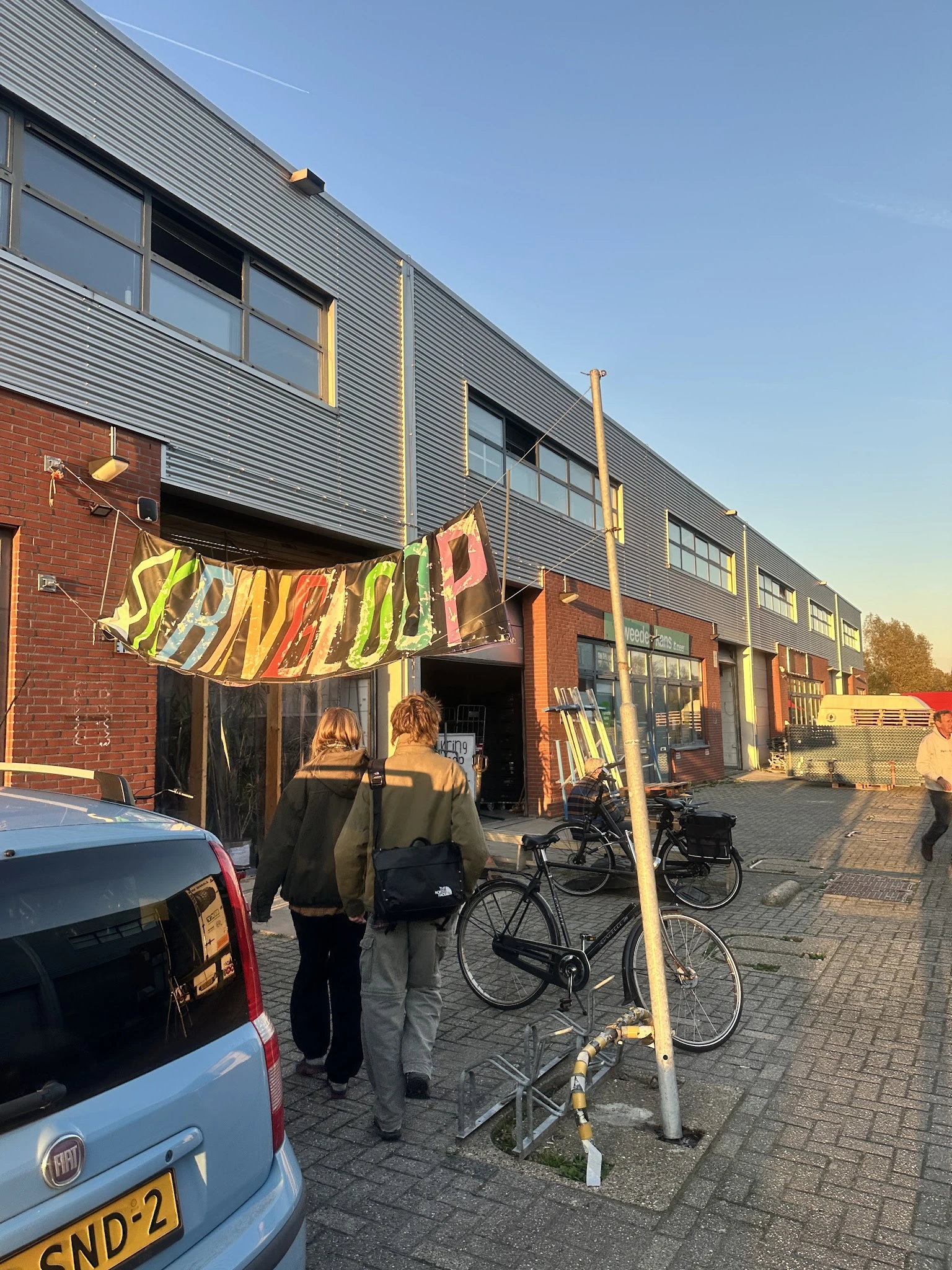Voorkant kringloopwinkel met geel spandoek, fietsen en persoon, bakstenen en metalen gevel