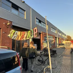 Voorkant kringloopwinkel met geel spandoek, fietsen en persoon, bakstenen en metalen gevel