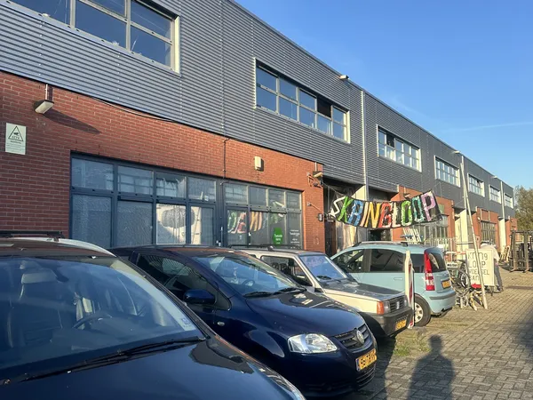 Gevel van kringloopwinkel met rode bakstenen en grijze metalen bovenbouw, grote ramen, uithanging met kleurig logo, parkeerpl
