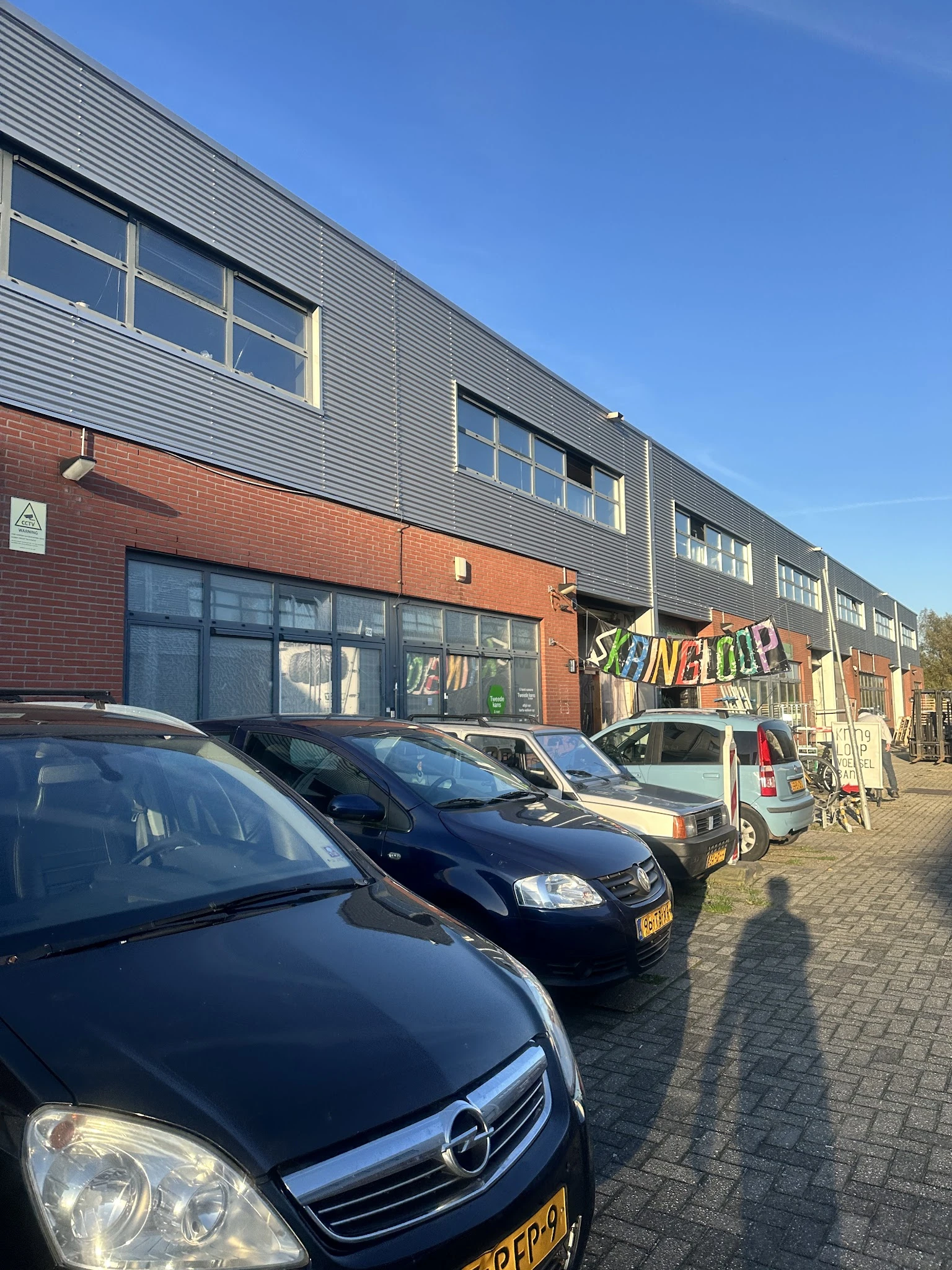 Gevel van kringloopwinkel met rode bakstenen en grijze metalen bovenbouw, grote ramen, uithanging met kleurig logo, parkeerpl