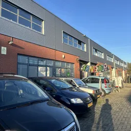 Gevel van kringloopwinkel met rode bakstenen en grijze metalen bovenbouw, grote ramen, uithanging met kleurig logo, parkeerpl