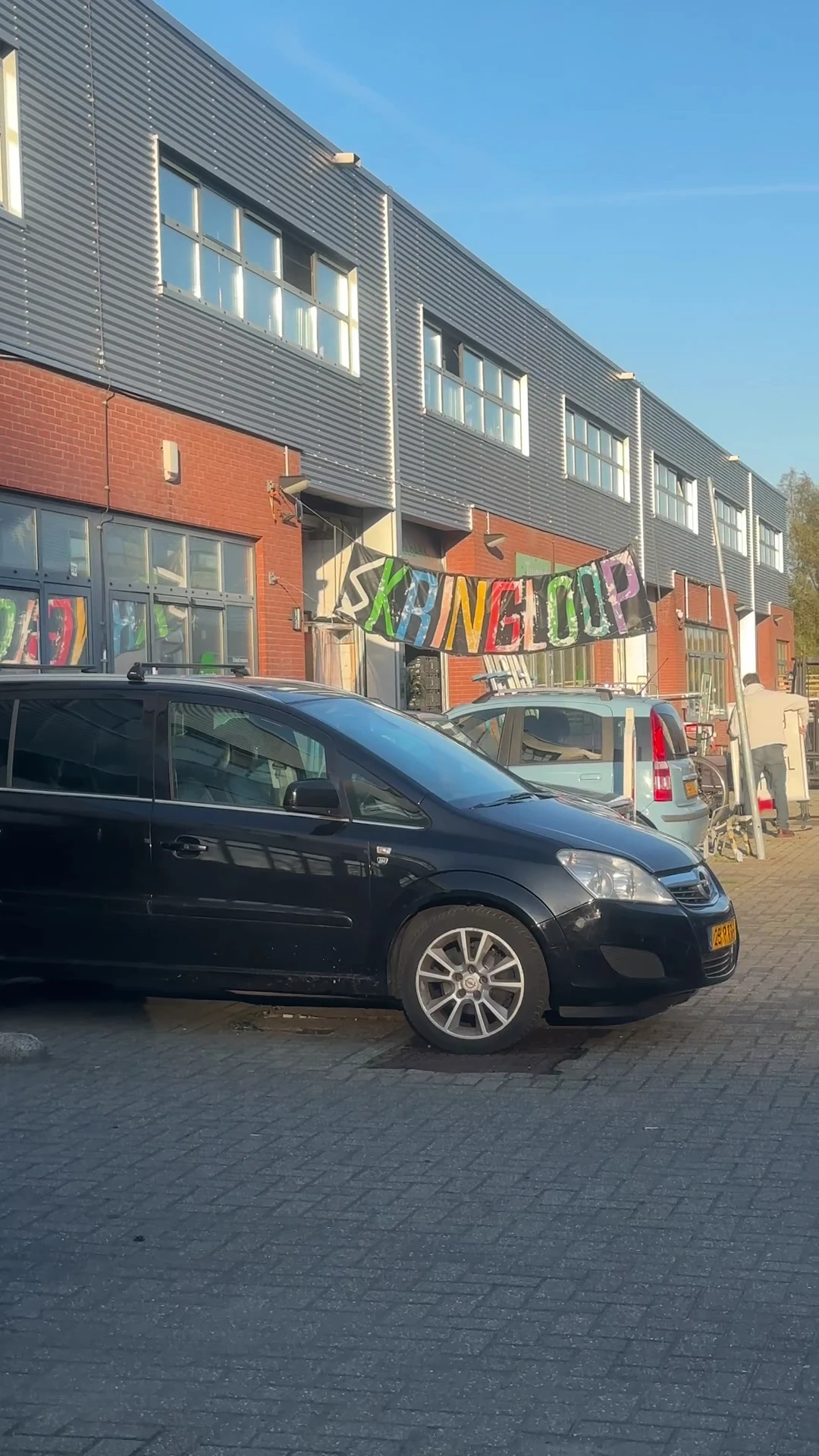 Voorkant van kringloopwinkel met zwarte auto geparkeerd ervoor en kleurrijk 'KRINGLOOP' spandoek boven de ingang