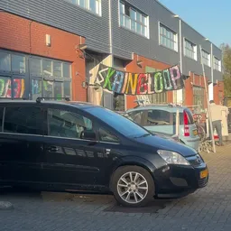 Voorkant van kringloopwinkel met zwarte auto geparkeerd ervoor en kleurrijk 'KRINGLOOP' spandoek boven de ingang