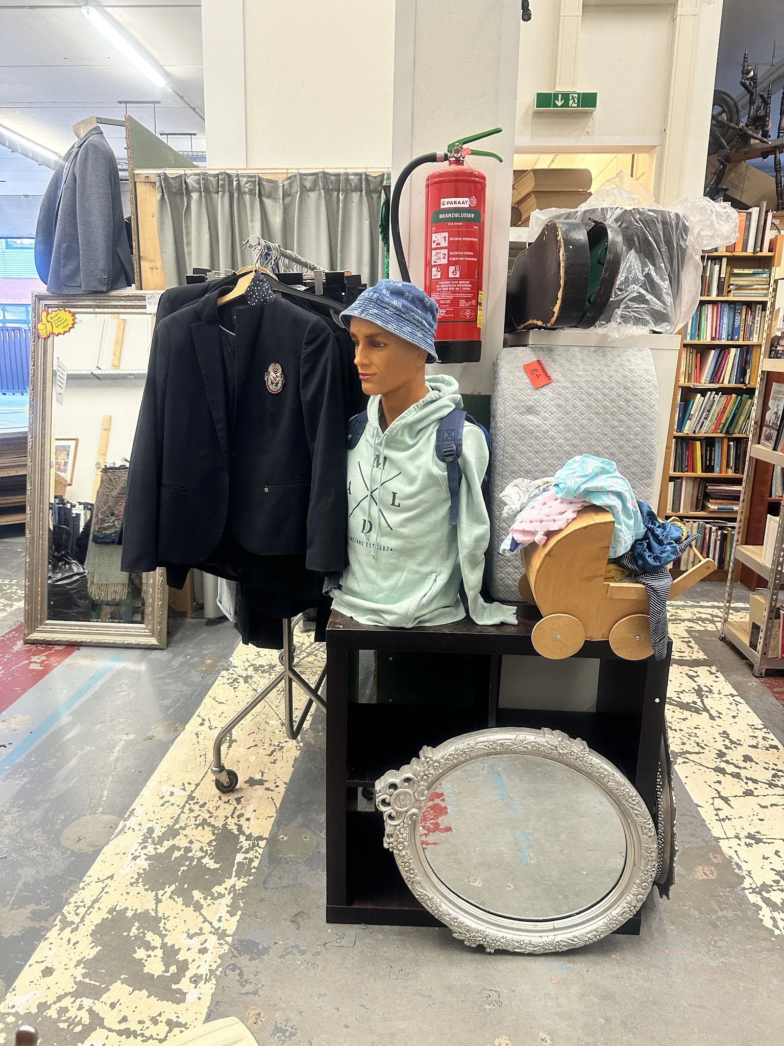 Winkelbinnenruimte met etalagepop gekleed in jasje en pet, omringd door kleding, boeken en huishoudartikelen