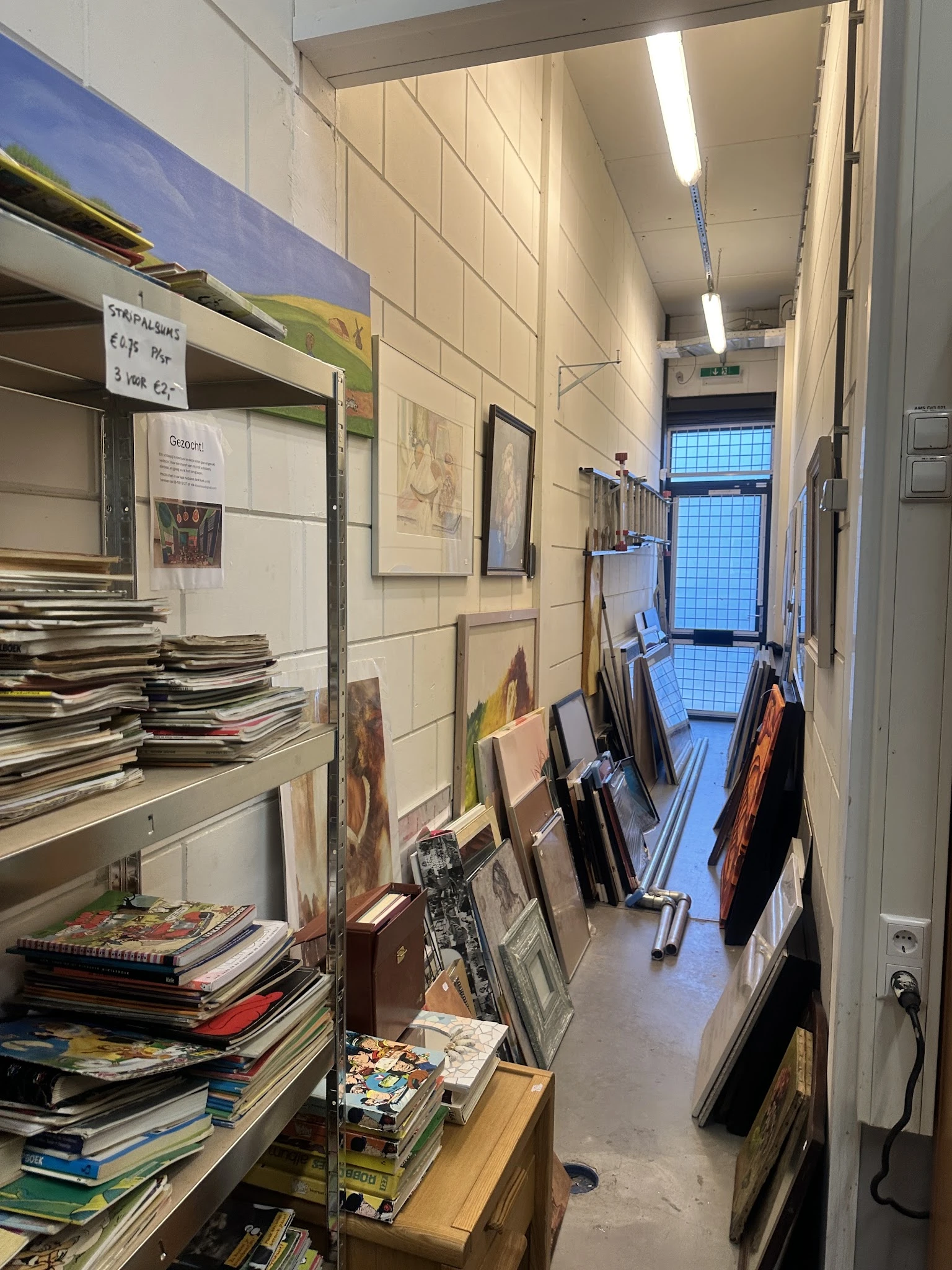 Nauwe gang in kringloopwinkel met aan wanden kunstwerken, tegen rechts muur diverse voorwerpen en tassen opgesteld