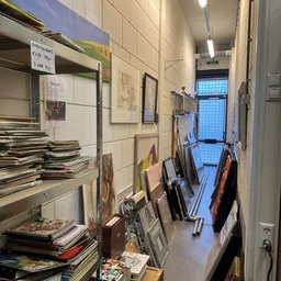 Nauwe gang in kringloopwinkel met aan wanden kunstwerken, tegen rechts muur diverse voorwerpen en tassen opgesteld
