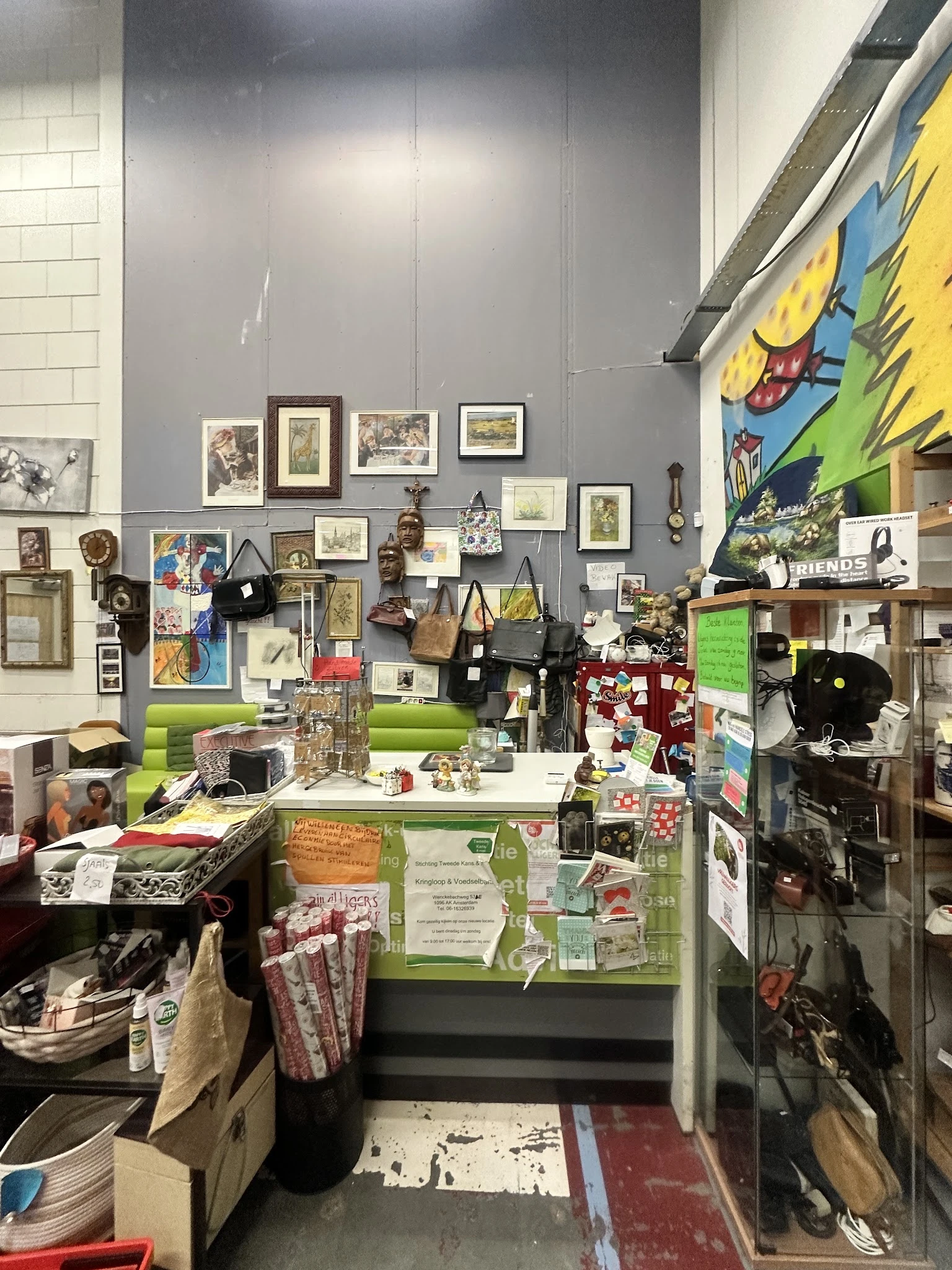 Winkelbinnenzicht met wanddecoratie, kunstwerk, boeken, huishoudartikelen en diverse tweedehands spullen op tafels en stellin