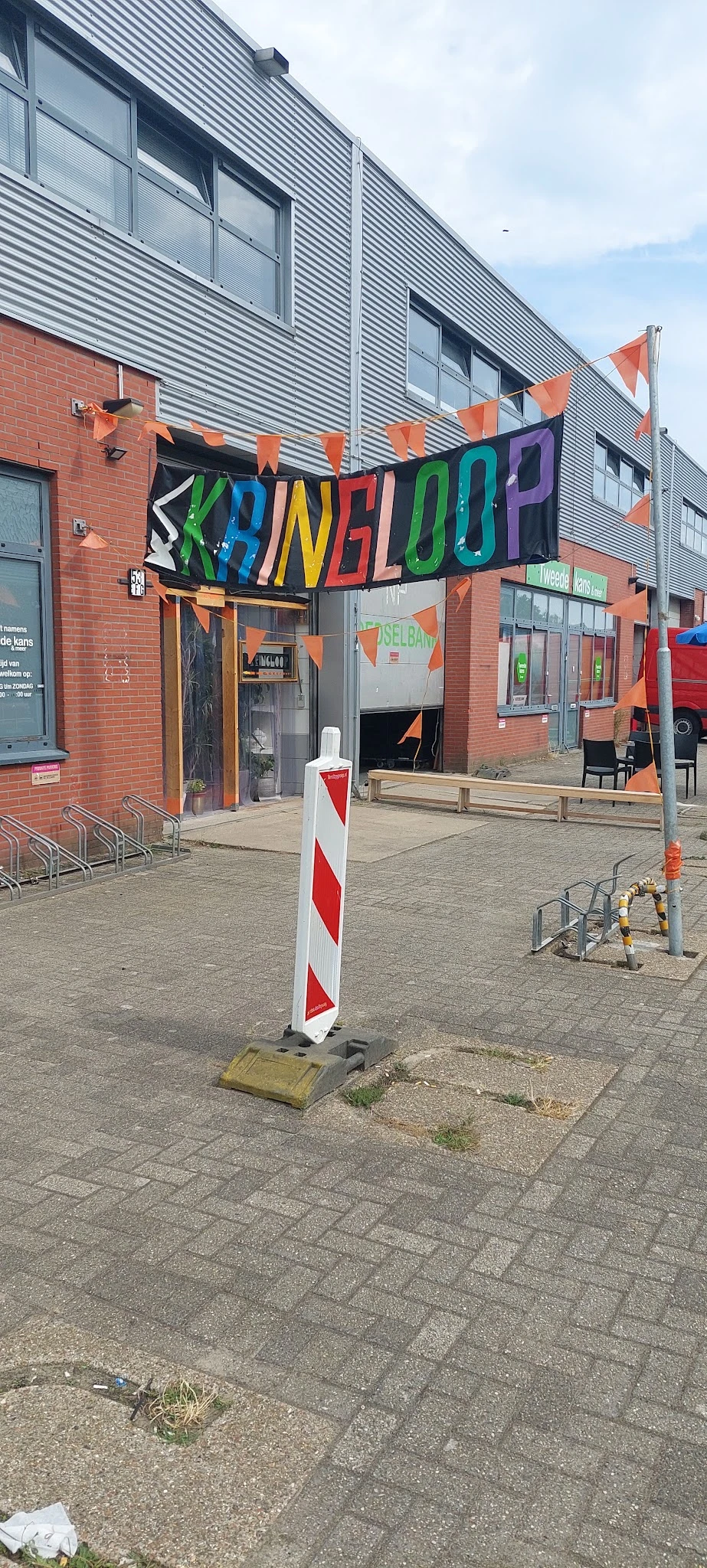 Gevel van kringloopwinkel met oranje en grijze kleuren, deels zichtbaar achter verkeerspoller en houten bankjes