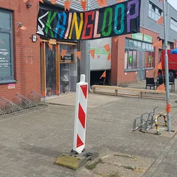 Gevel van kringloopwinkel met oranje en grijze kleuren, deels zichtbaar achter verkeerspoller en houten bankjes