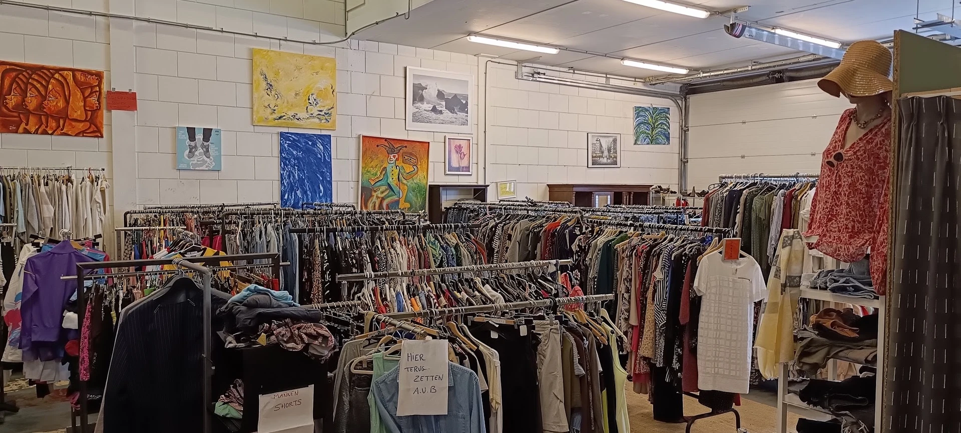 Rekken met tweedehands kleding en kunstwerken aan de muur in een kringloopwinkel