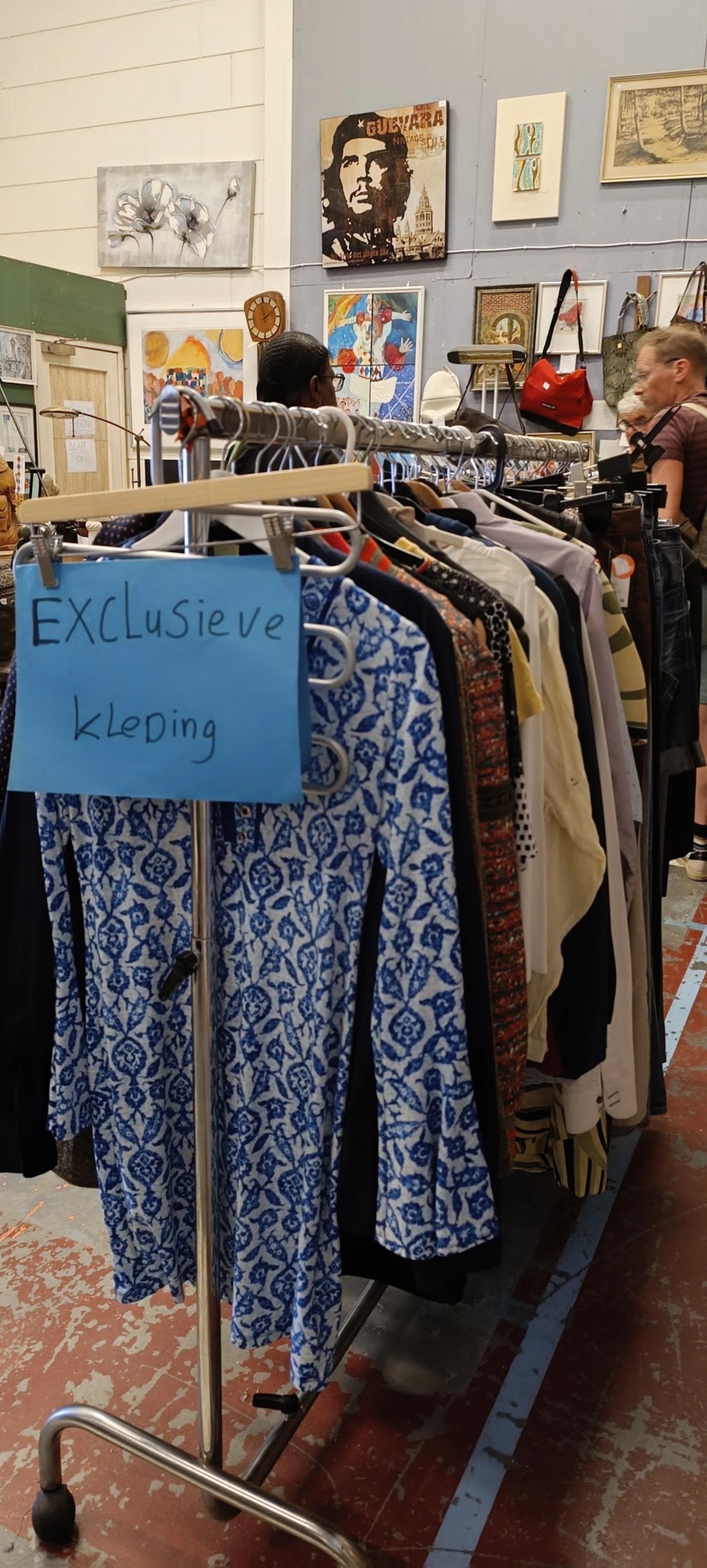 Rekken met tweedehands kleding, waaronder een blauw-wit gemusterd kledingstuk met een bordje 'EXCLUSIEVE kleping'