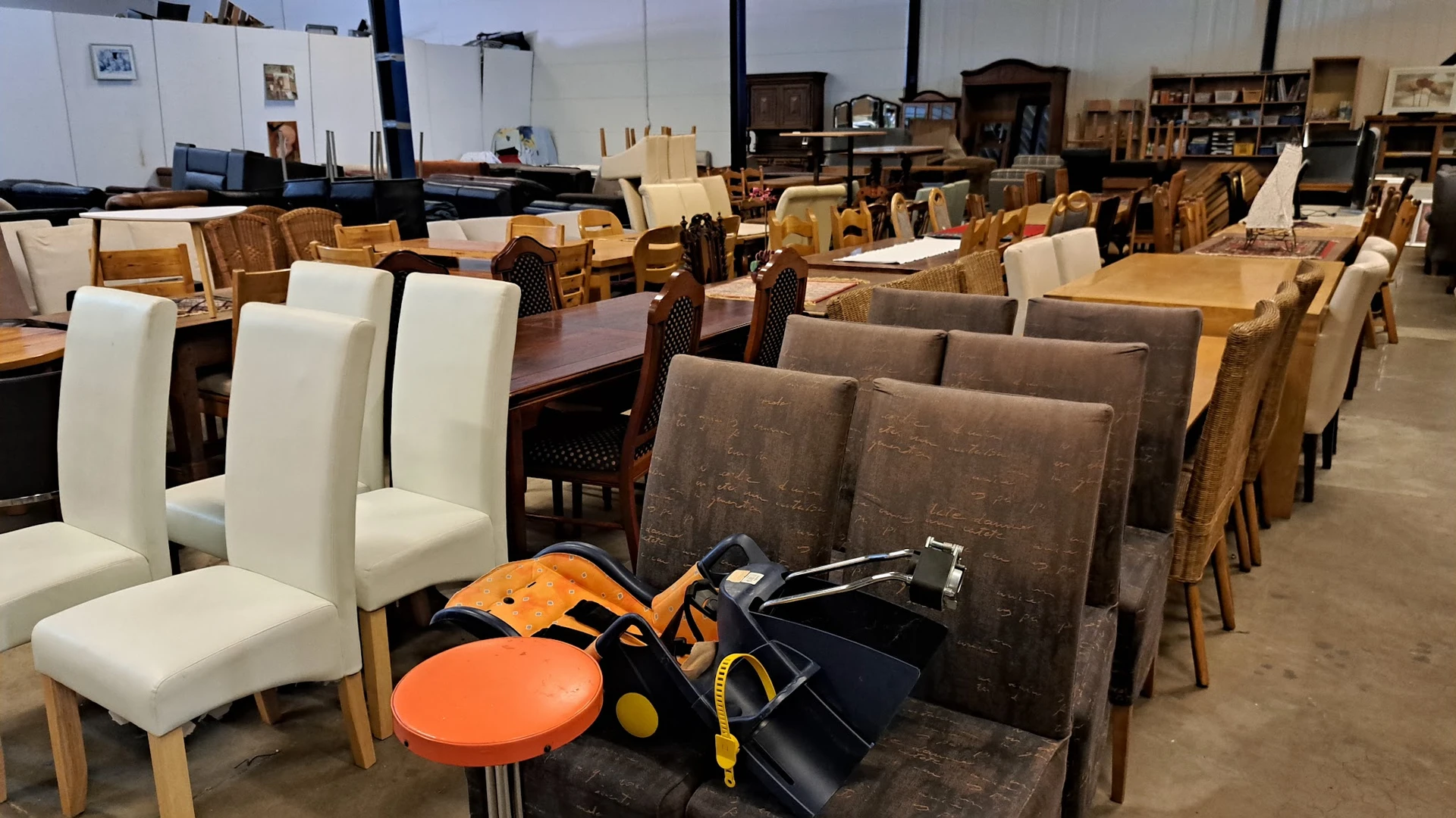 Diverse meubels en huishoudartikelen in een kringloopwinkel: stoelen, tafels, een oranje krukje en andere spullen