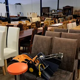 Diverse meubels en huishoudartikelen in een kringloopwinkel: stoelen, tafels, een oranje krukje en andere spullen