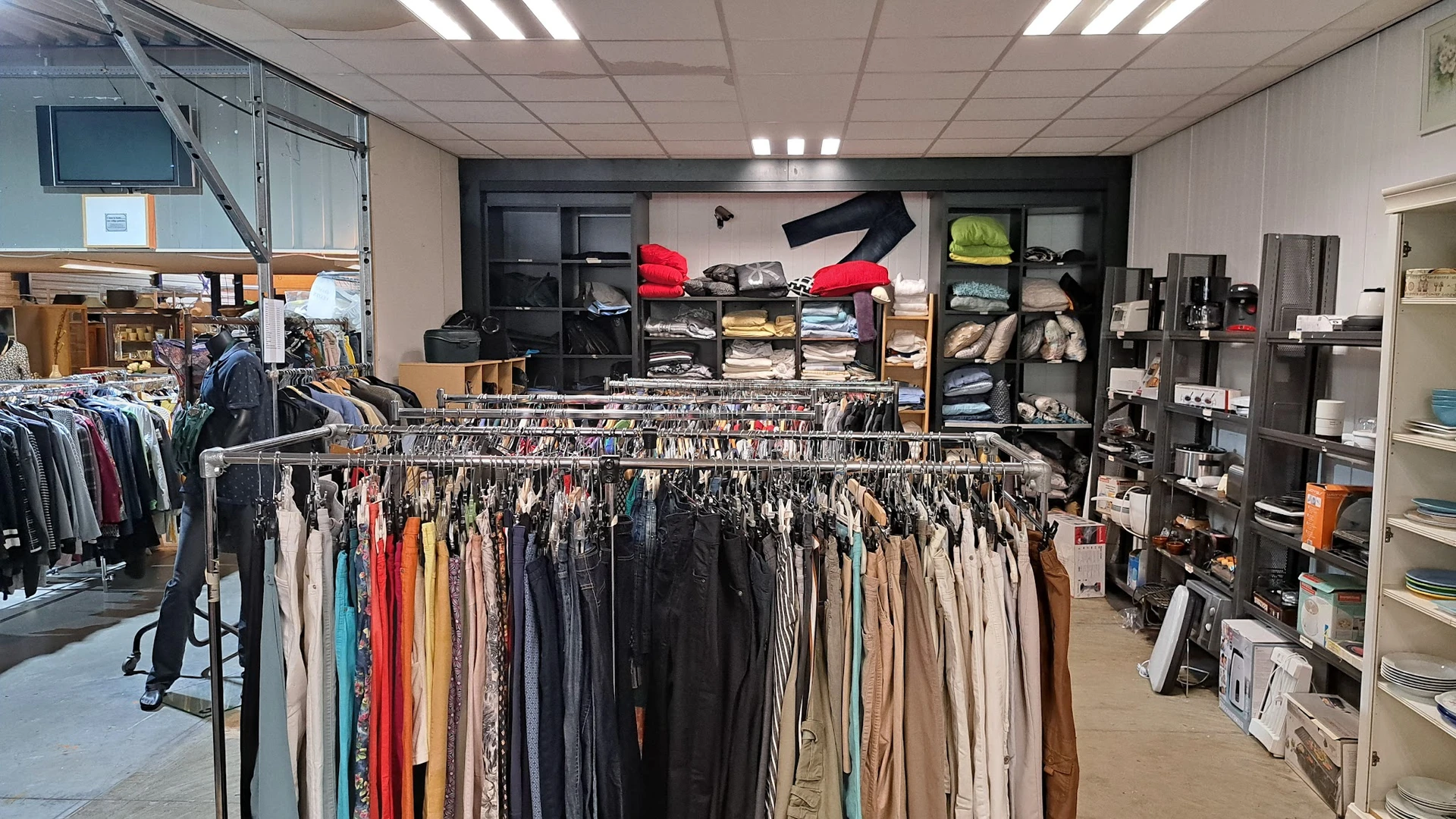 Rekken met tweedehands kleding in diverse kleuren, op de achtergrond stellingen met dozen en tassen