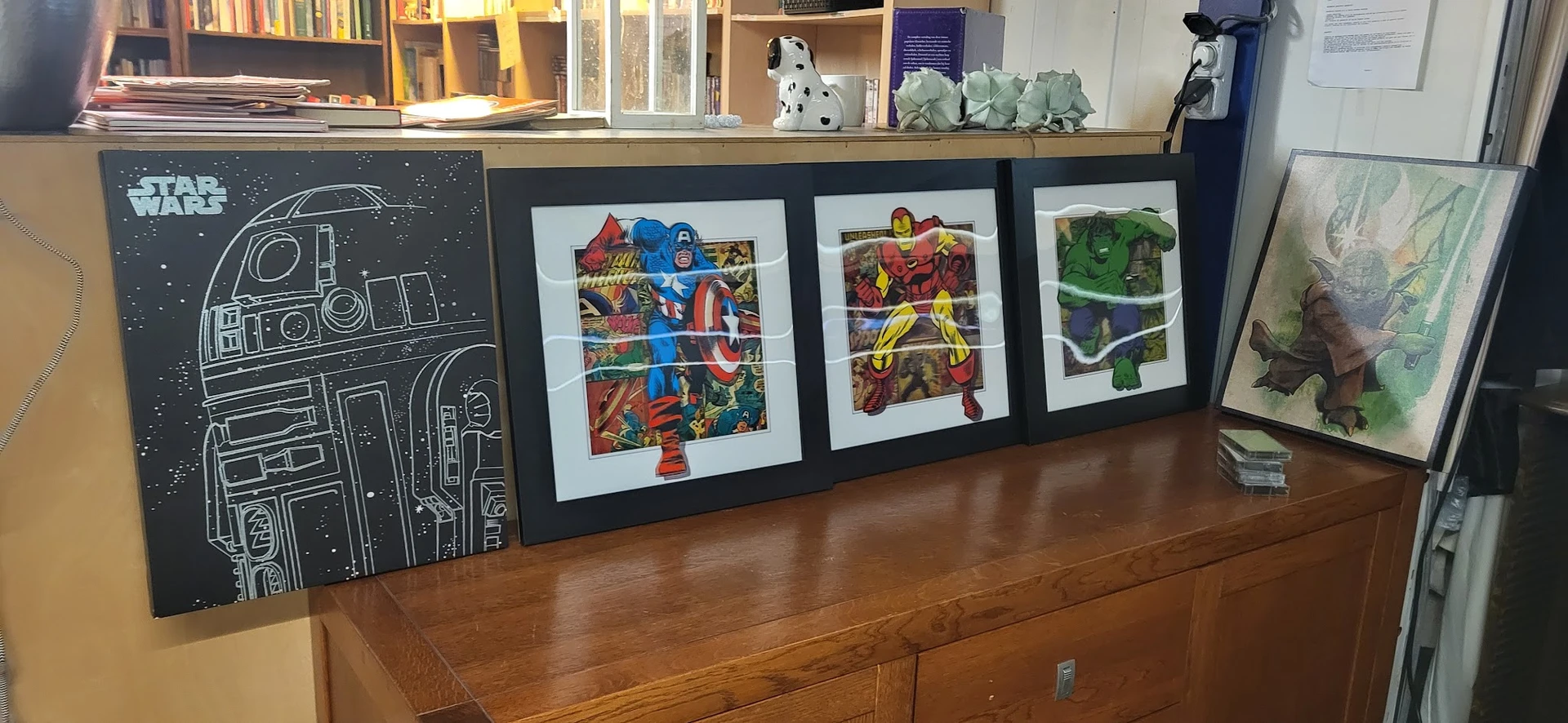 Drie ingelijste posters met superhelden afbeeldingen op houten dressoir, bovenop boeken en decoratie