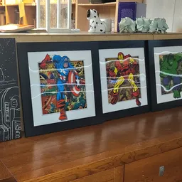 Drie ingelijste posters met superhelden afbeeldingen op houten dressoir, bovenop boeken en decoratie