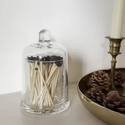 Glazen pot met houten luciferstokjes met zwarte koppen naast een koperen kandelaar met dennenappels