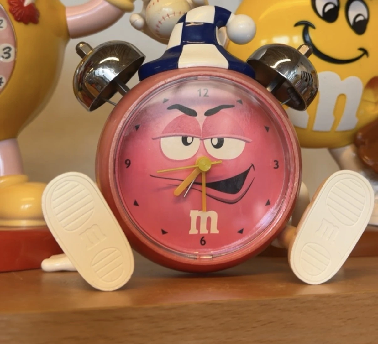 Roze M&M's wekker met bruine oren, witte voeten en smilend gezicht op houten oppervlak