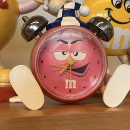 Roze M&M's wekker met bruine oren, witte voeten en smilend gezicht op houten oppervlak
